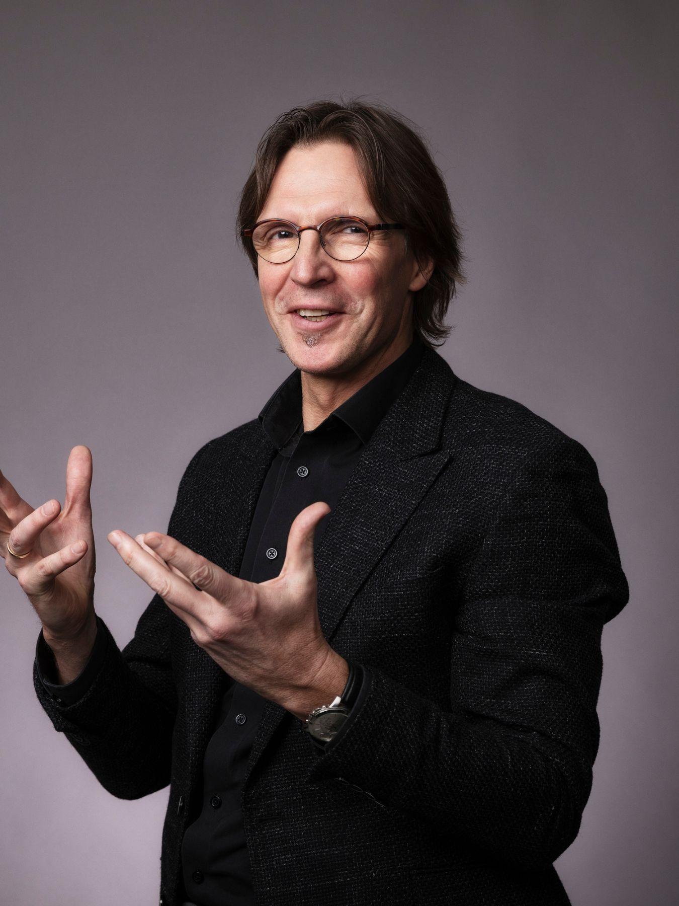 Putniņš wears round glasses and smiles. He gesticulates as if he is explaining something.