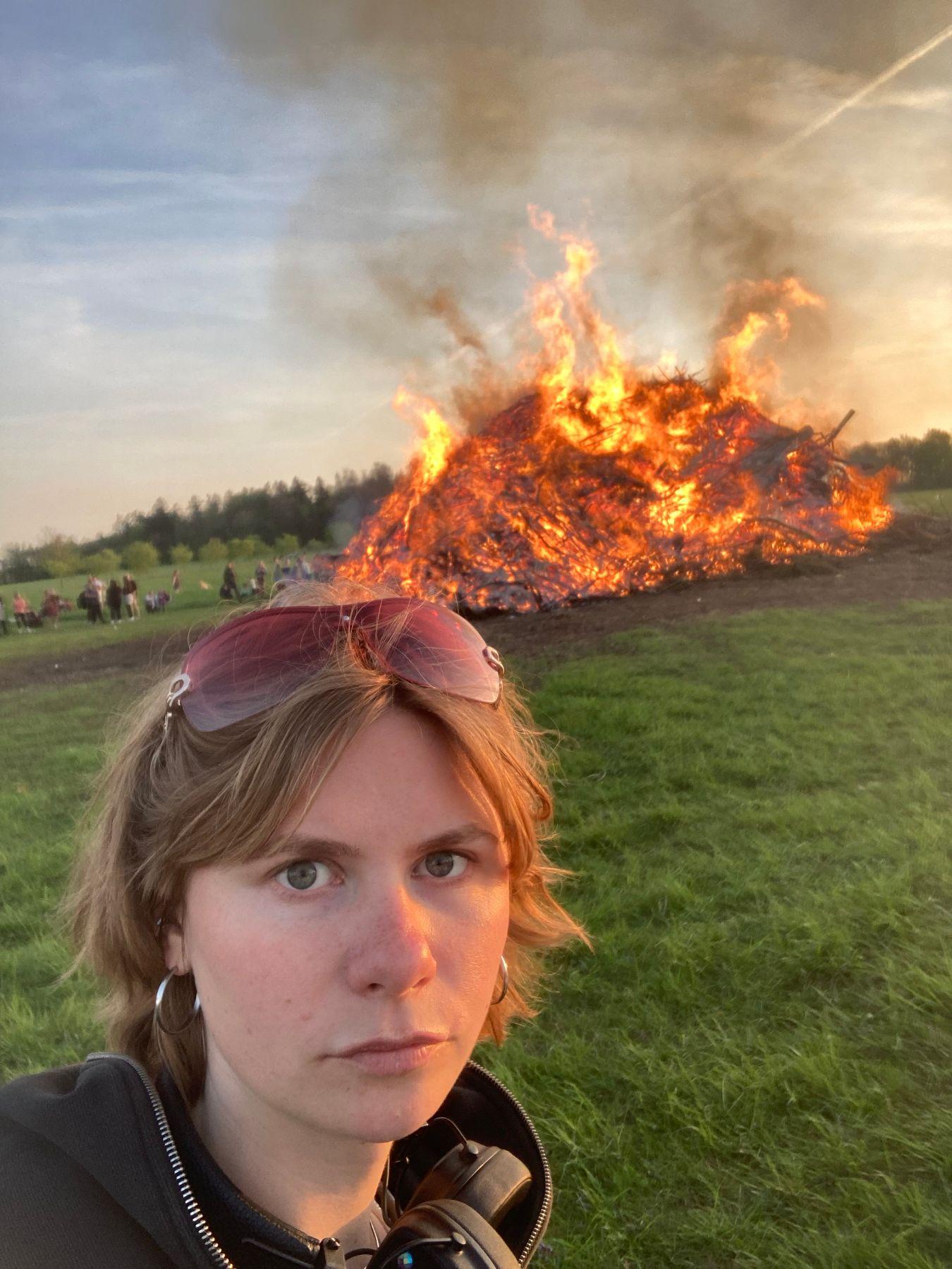 A woman with short blonde hair and sunglasses on her head standing in front of a burning pile of rubbish