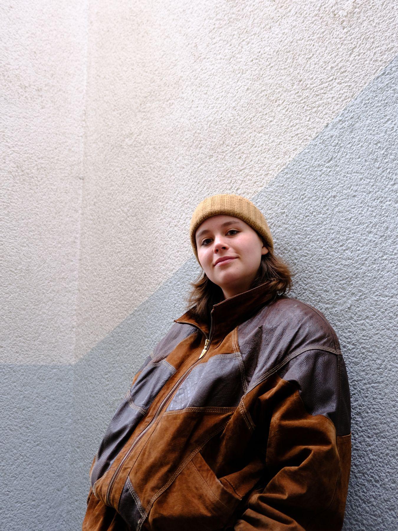 Das Bild zeigt eine junge Frau, die vor einer Wand steht. Die Person trägt eine braune Jacke mit schwarzen Akzenten und eine Strickmütze. Die Wand hinter der Person hat eine strukturierte Oberfläche und ist in einem neutralen Farbton gehalten. Das Licht scheint von oben zu kommen und wirft Schatten auf die Wand und die Person.
