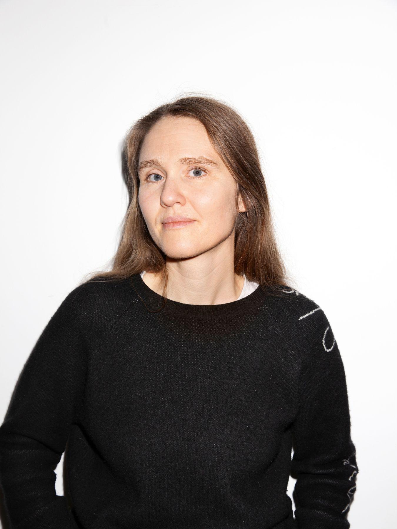 Person in dunklem Pullover steht vor weißer Wand, Hände in den Taschen, Oberkörper frontal.