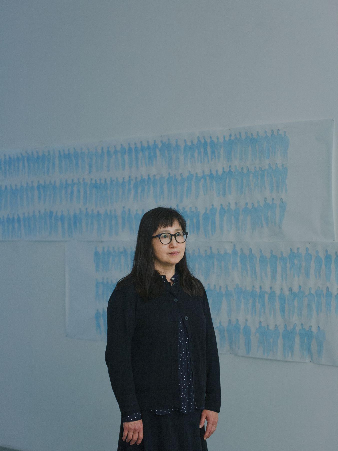 Don Mee Choi steht vor einer Malerei, die zahlreiche Konturen von Personen zeigt.