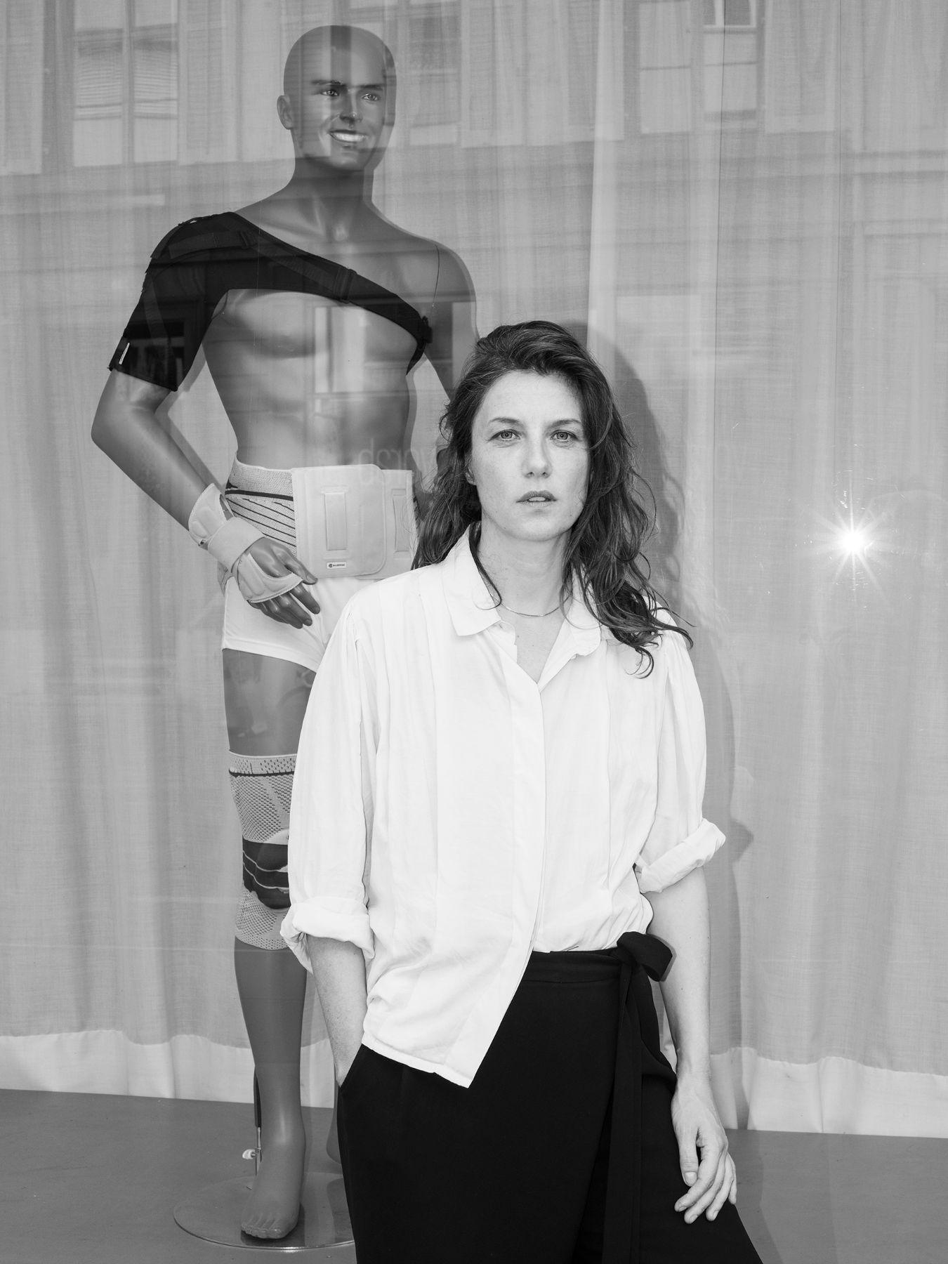 Eine Frau mit schulterlangen dunklen Haaren steht mit einer Hand in der Hosentasche vor einer männlichen Schaufensterpuppe.