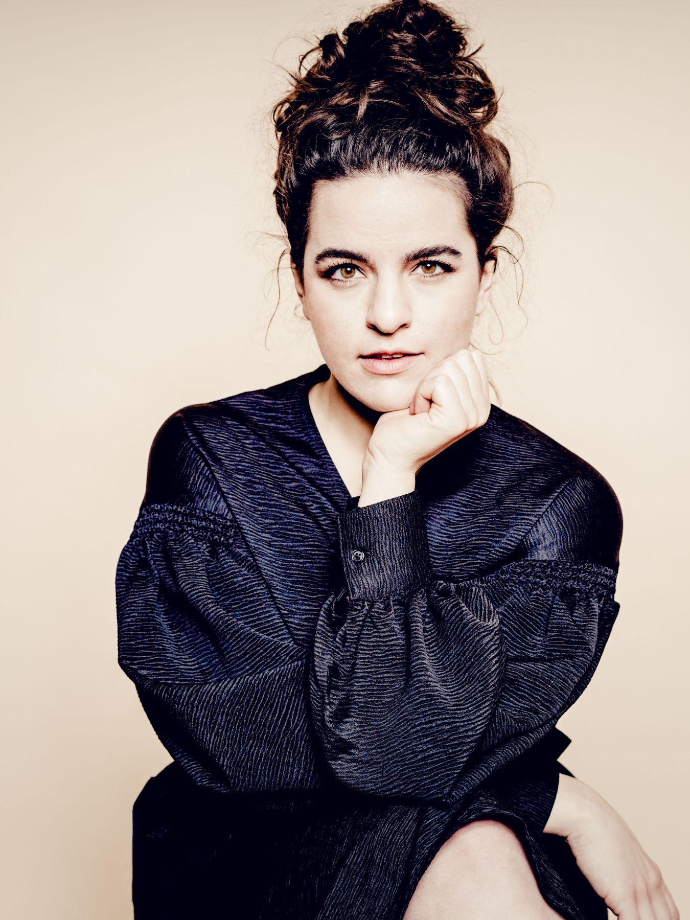 A woman sits cross-legged, crosses her arms and looks into the camera.