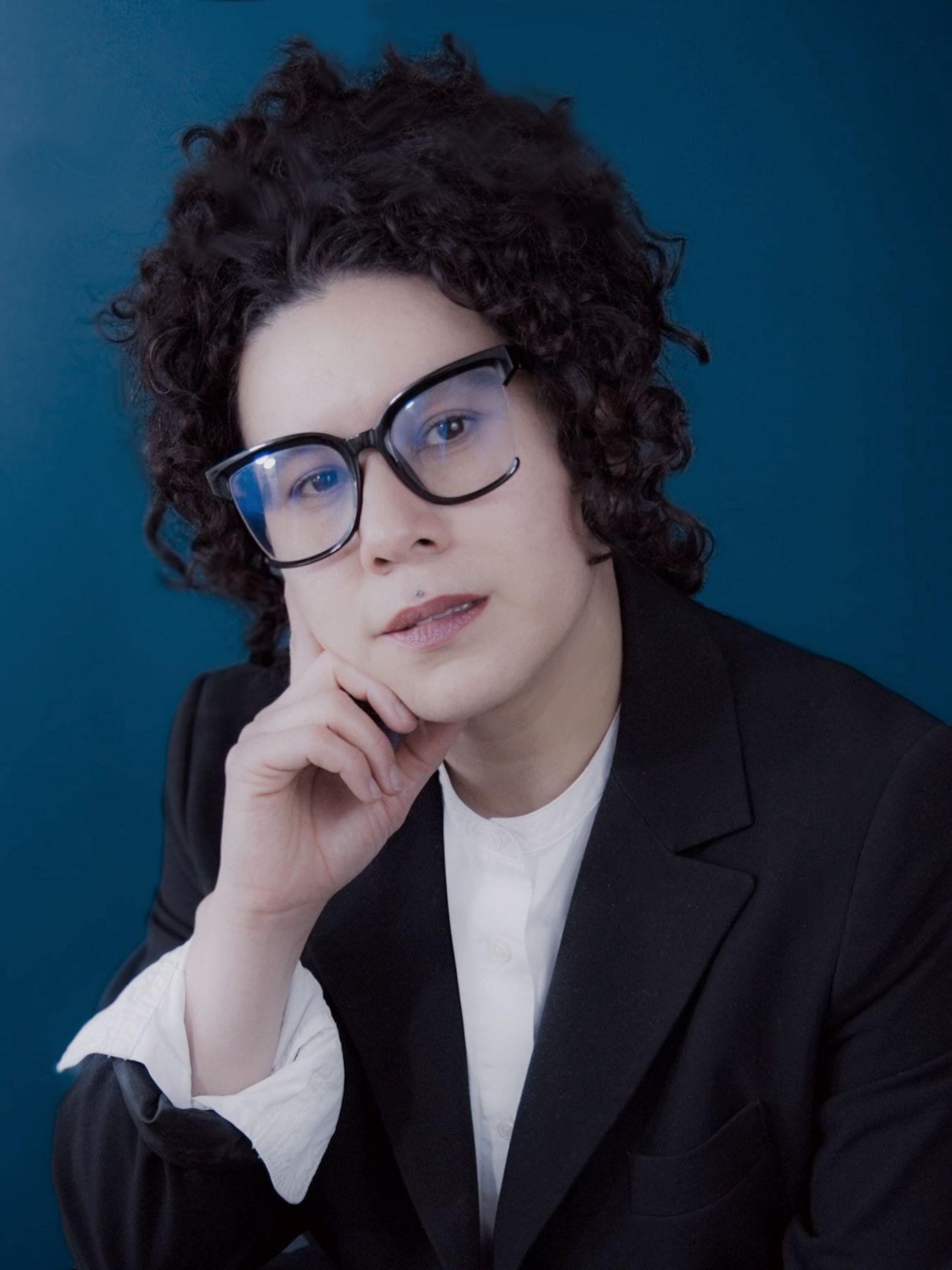 Eine Frau mit Brille, schwarzen, lockigen Haaren, in Hemd und Sakko.