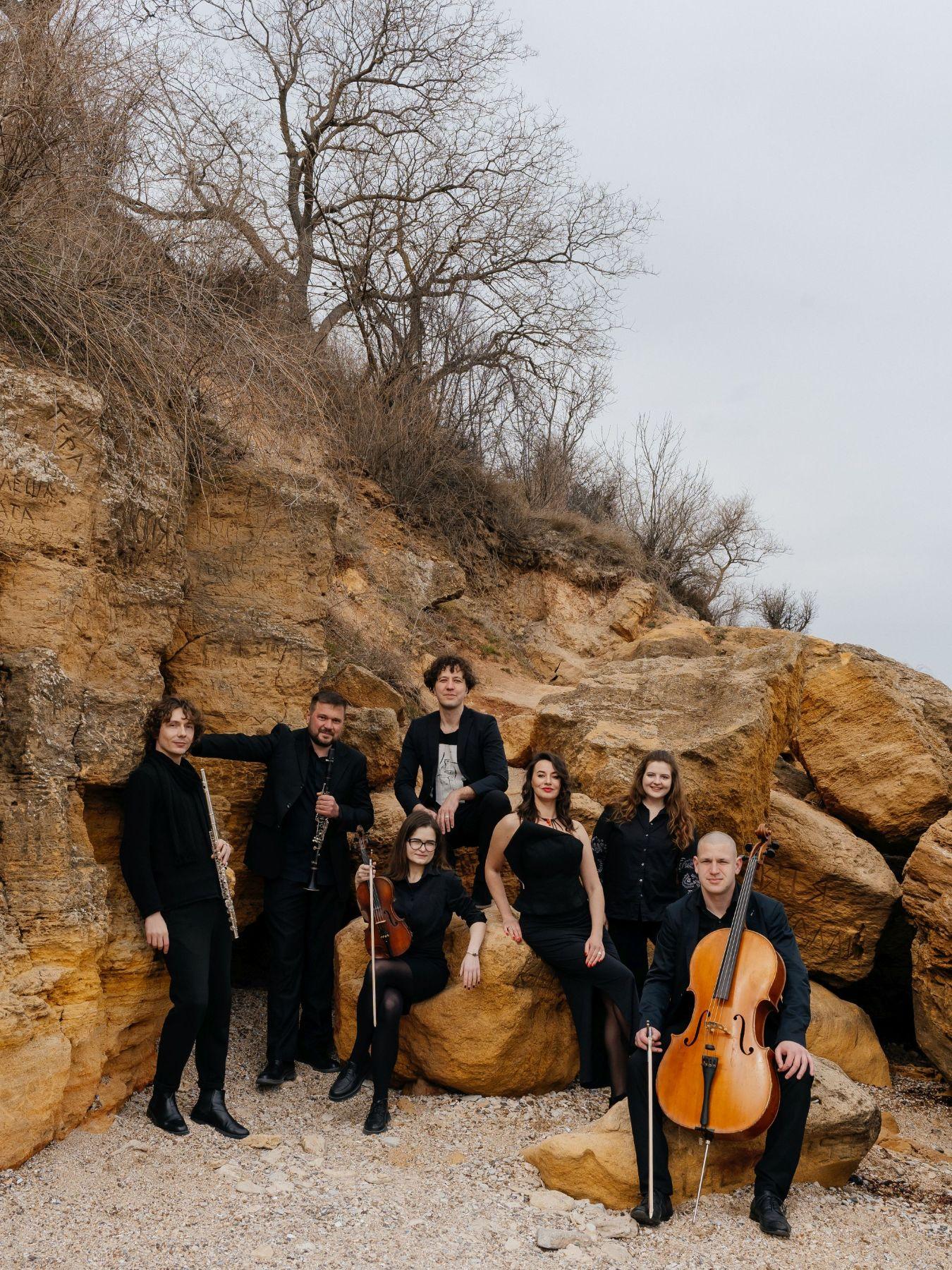 Menschen in Anzügen und Abendkleidern vor einem Felsen, ein paar halten Instrumente.