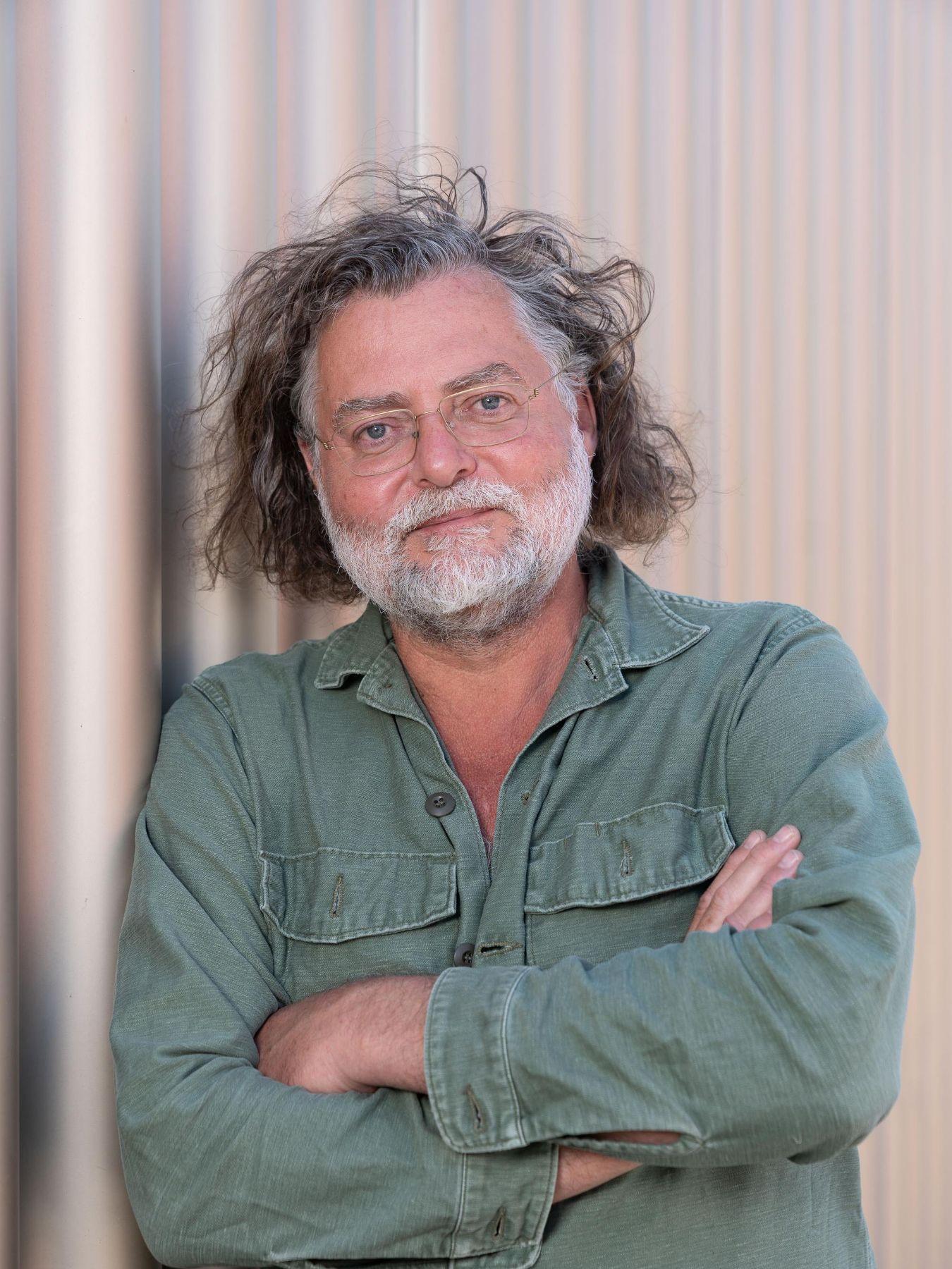 A person with gray, shoulder‑length curls and a green shirt leans against a light corrugated‑metal wall and looks neutrally into the camera.
