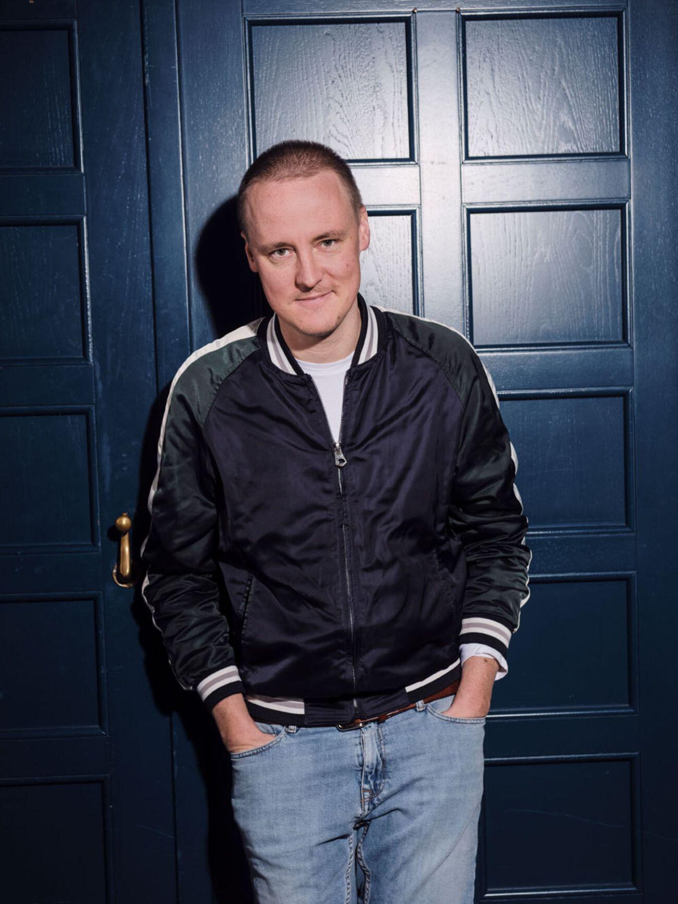 A person stands in front of blue paneled doors, wearing a black bomber jacket and light blue jeans, with hands in pockets, facing the camera.