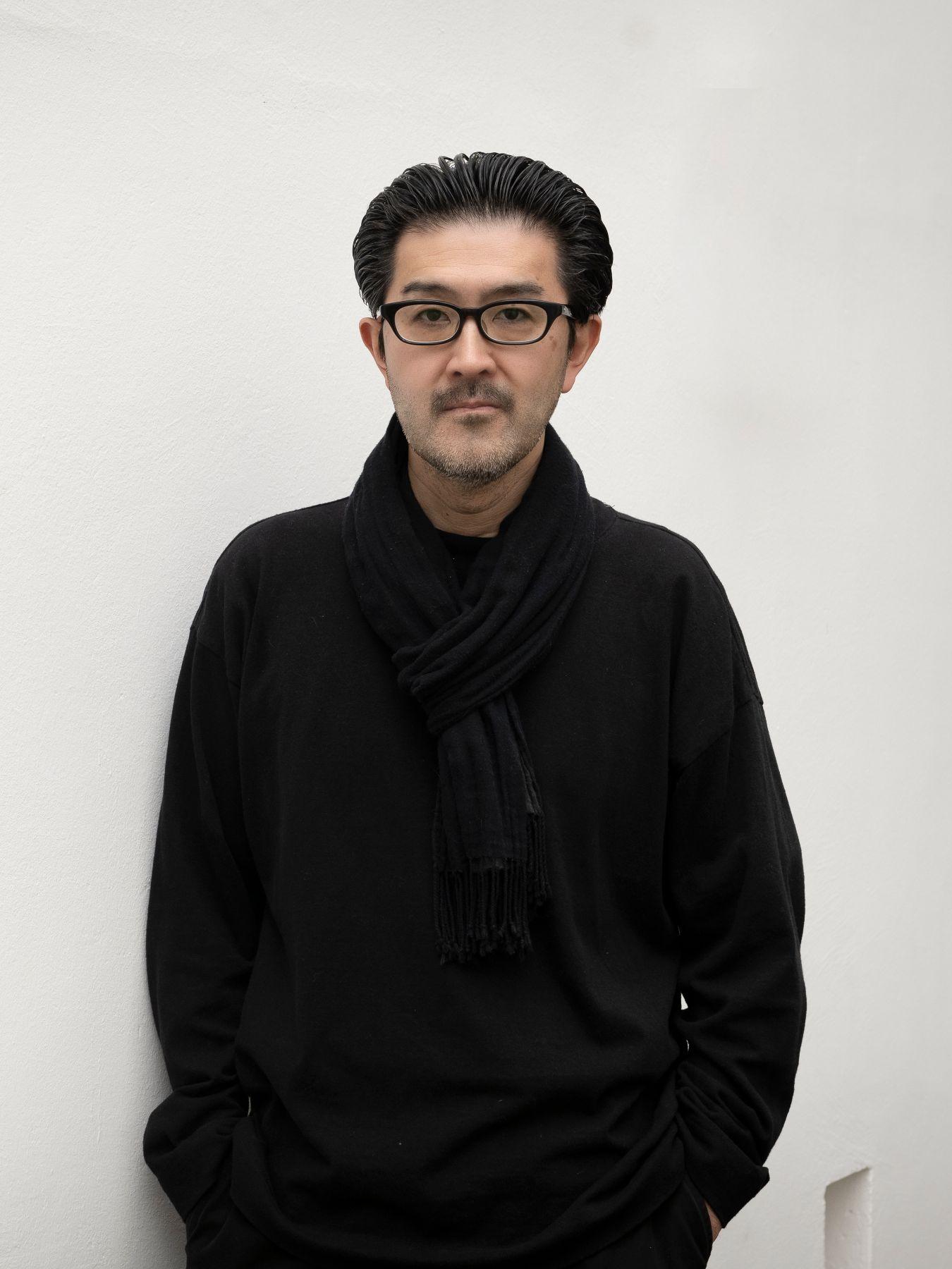 Person dressed in dark clothing standing against a plain light-coloured wall with hands in pockets.