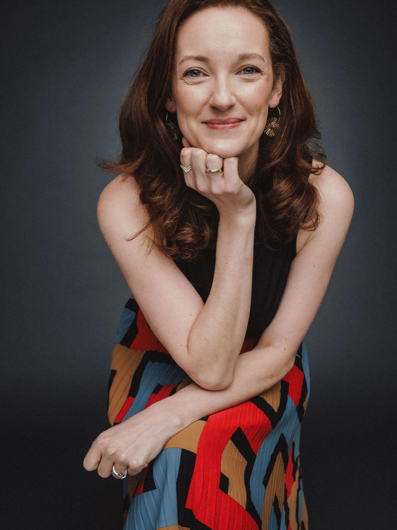 Eine Frau mit langen braunen Haaren, ihren Kopf auf die Hände gestützt, schaut in die Kamera. Sie trägt einen farbigen langen Rock.