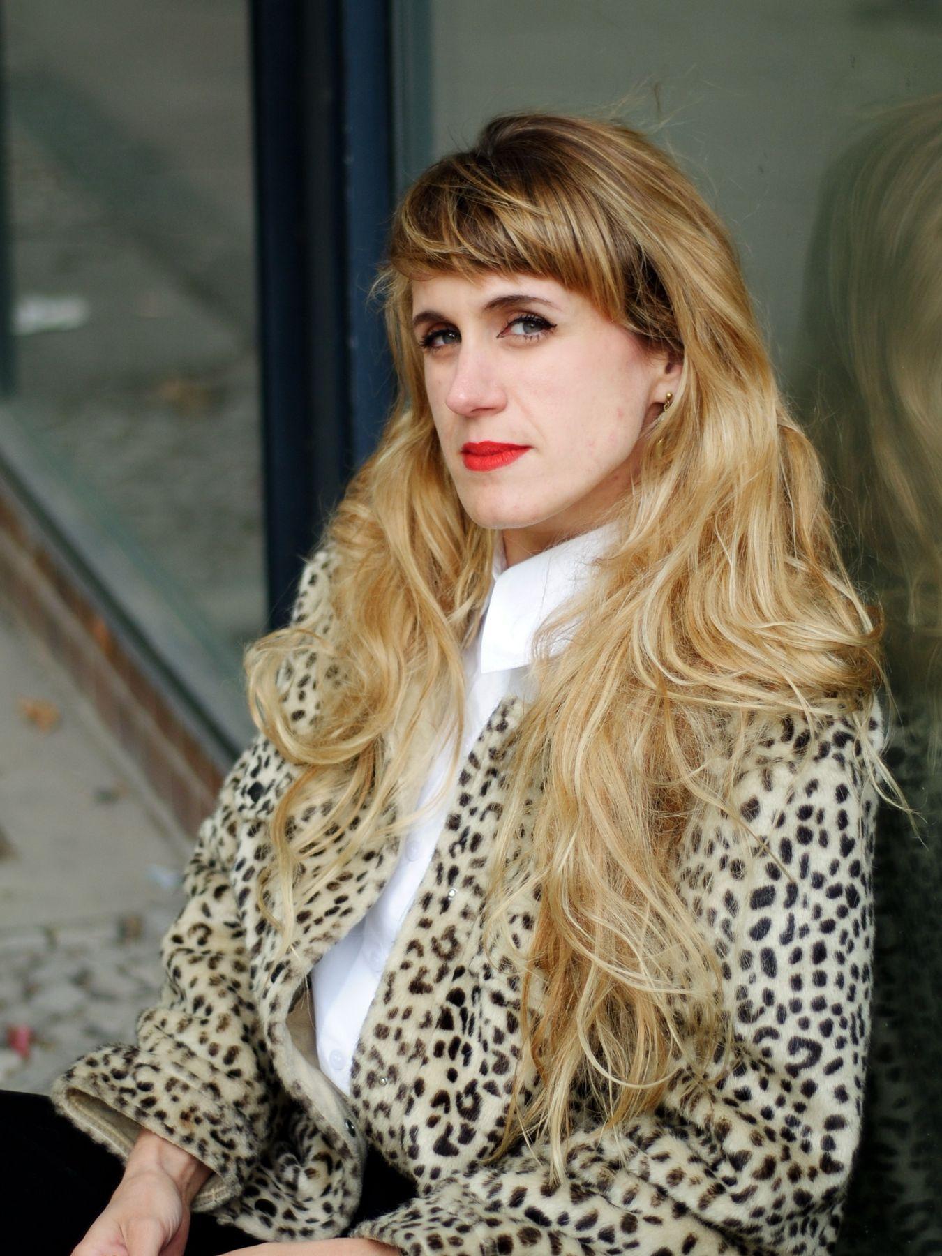A woman with long blonde hair, red lipstick and a leopard-print jacket looking sceptically into the camera