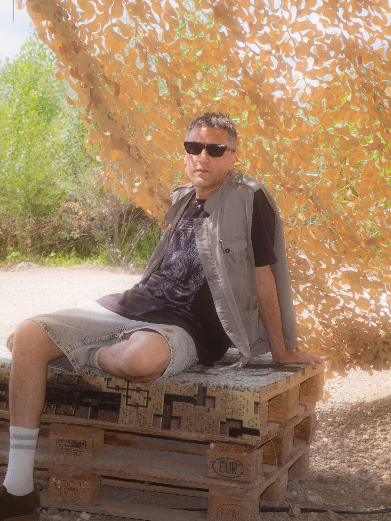 Ein Mann mit einer Sonnenbrille und kurzen Haaren sitzt lässig auf zwei Holzpaletten, dei Sonne scheint und hinter ihm sieht man grüne Büsche und ein Netz, das Schatten spendet.
