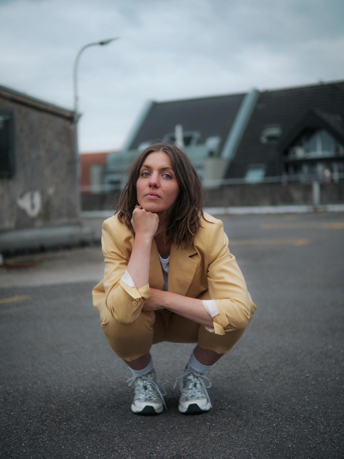 Eine junge Frau sitzt in der Hocke und stützt ihren Kopf auf ihrer Hand ab. Sie trägt einen gelben Anzug und blickt in die Kamera.