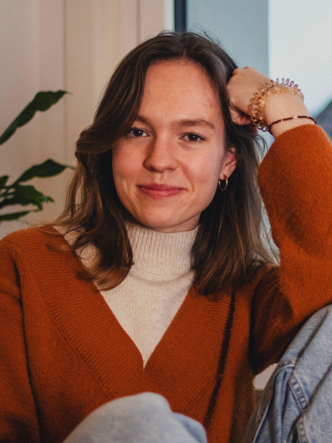 Das Bild zeigt eine junge Frau mit braunen Haaren, die lächelt und in die Kamera schaut. Sie trägt einen orangefarbenen Pullover über einem Rollkragenpullover und Jeans. Sie hat eine Hand am Kopf und trägt einige Armbänder. Im Hintergrund sind ein verschwommener Teil einer Zimmerpflanze und eine Wand zu sehen. Das Foto ist gut beleuchtet und hat einen warmen Ton.