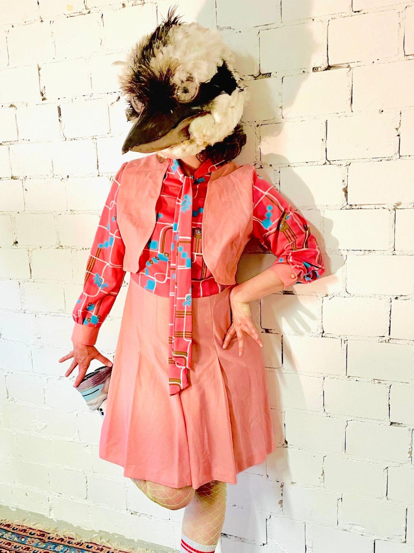 Foto einer Person in einem rosa Outfit. Sie trägt eine Vogelmaske und hebt ein Bein nach hinten an.
