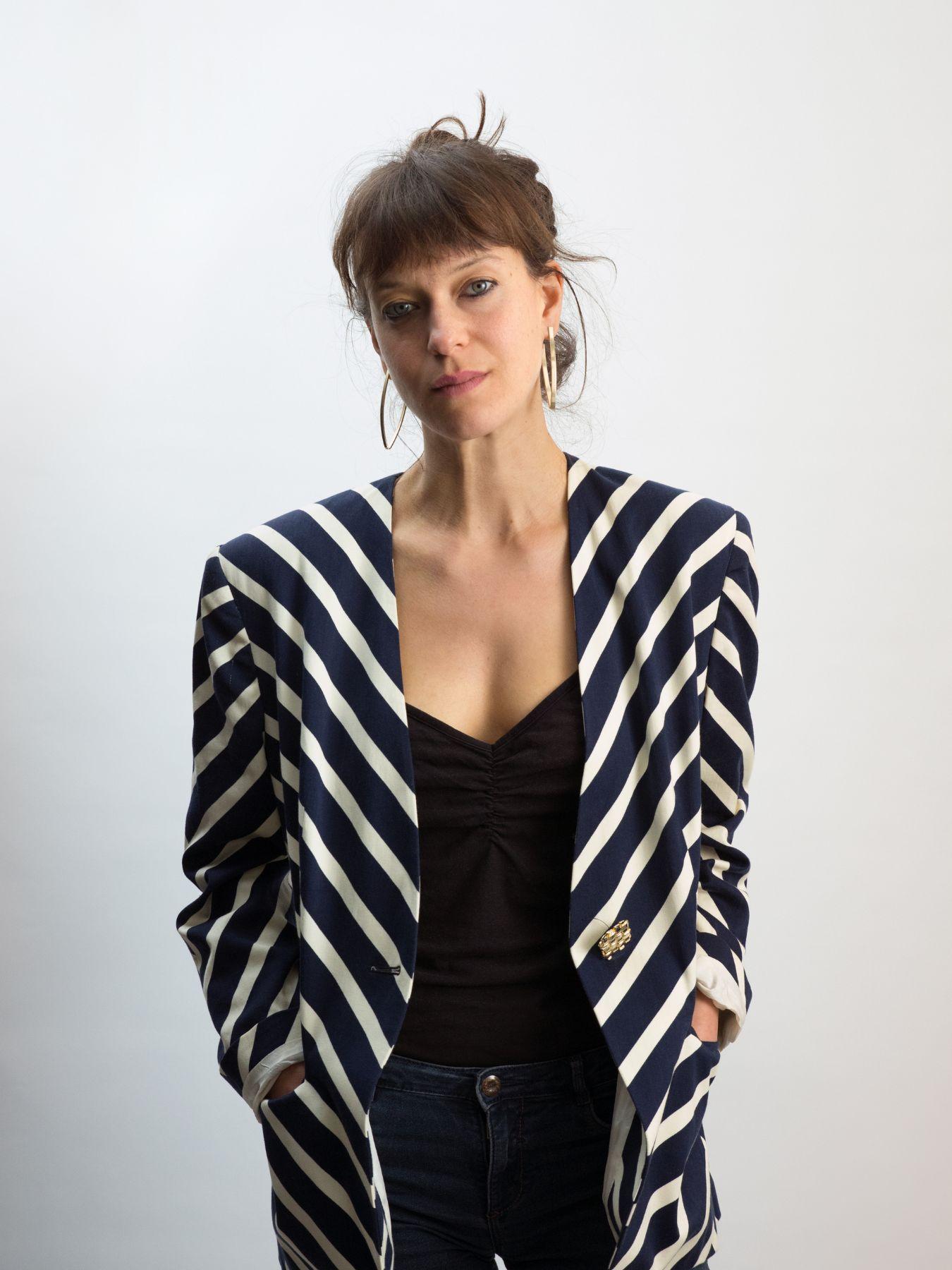 Halbporträt einer Person mit langen braunen Haaren im Zopf, trägt einen gestreiften Blazer.