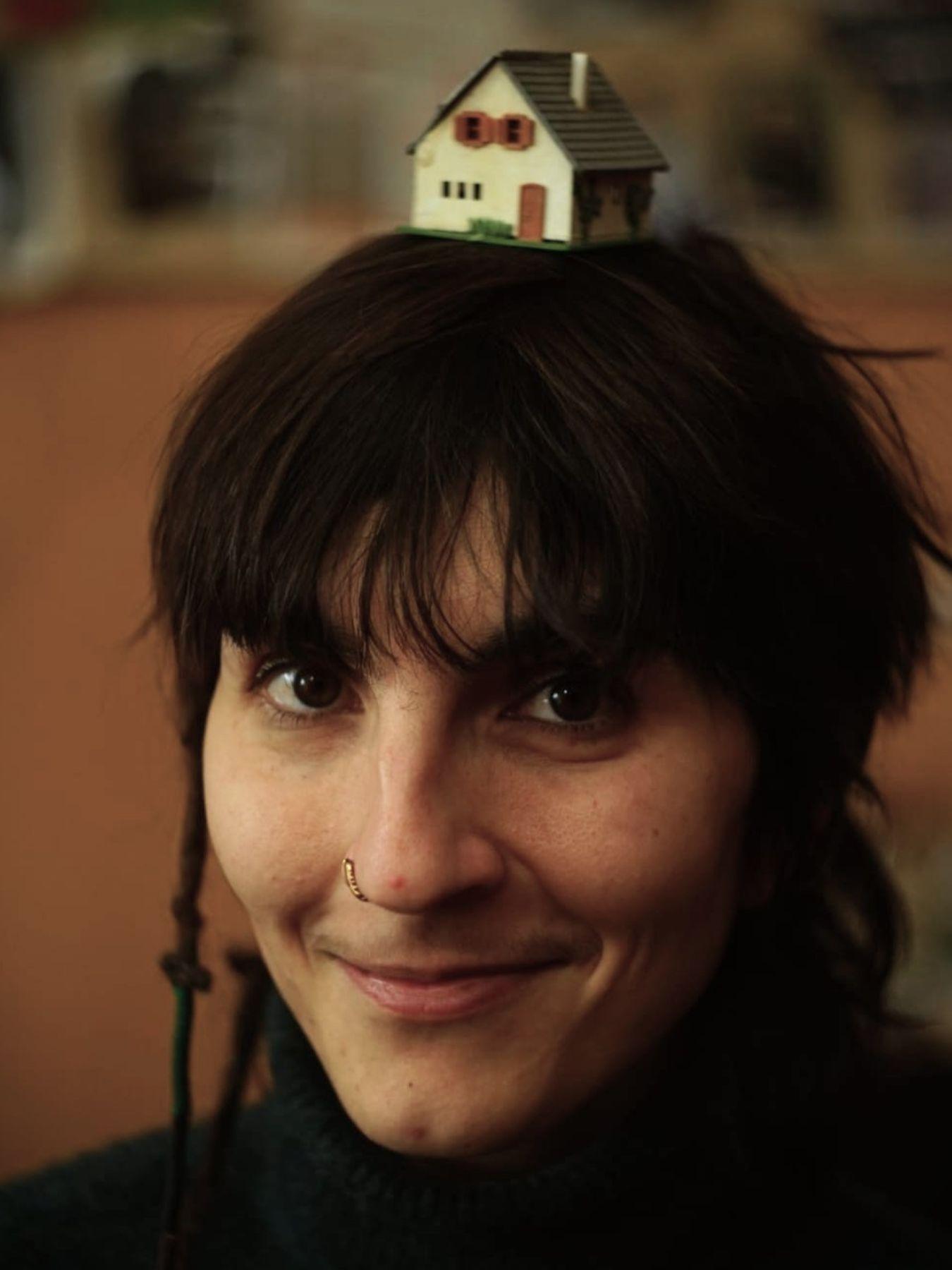 Eine Person mit Nasenpiercing und dunklen Haaren, die ein Miniaturhaus auf dem Kopf trägt
