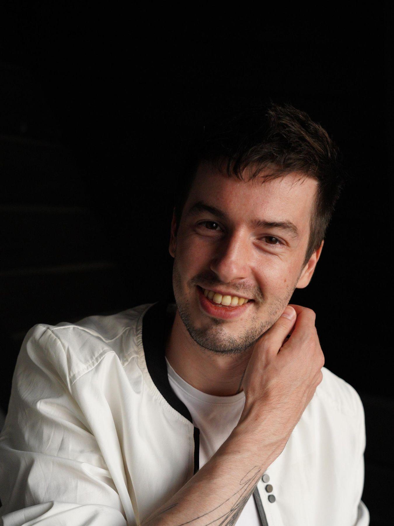 Ein junger Mann mit kurzen Haaren in einem weißen Sweatshirt und einem Tattoo auf dem Unterarm.