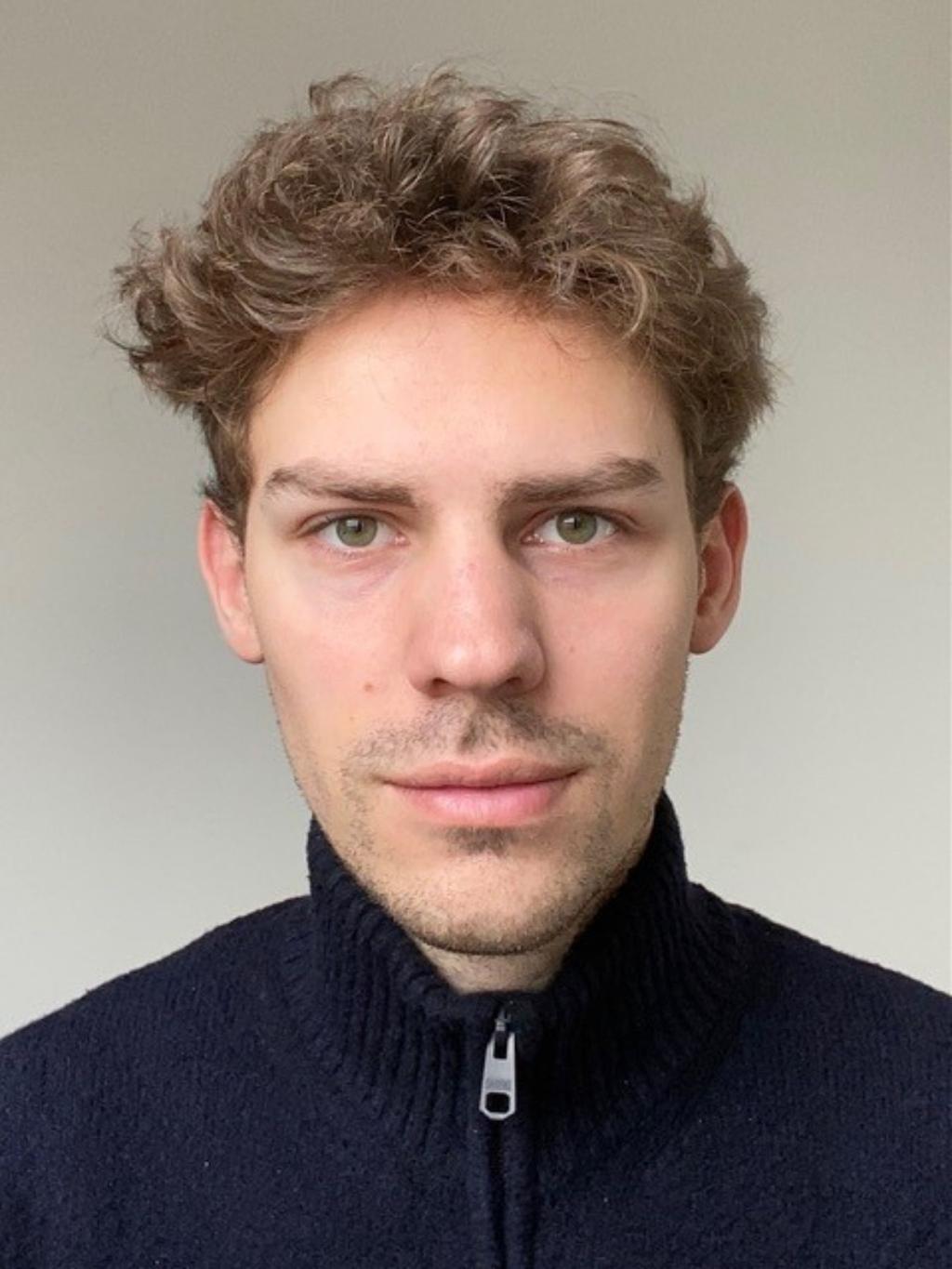 Das Bild zeigt ein Porträt eines jungen Mannes mit hellbraunem, gewelltem Haar. Er hat helle Haut und trägt einen dunkelblauen Pullover mit Reißverschluss. Der Hintergrund ist einfarbig hellgrau oder weiß.