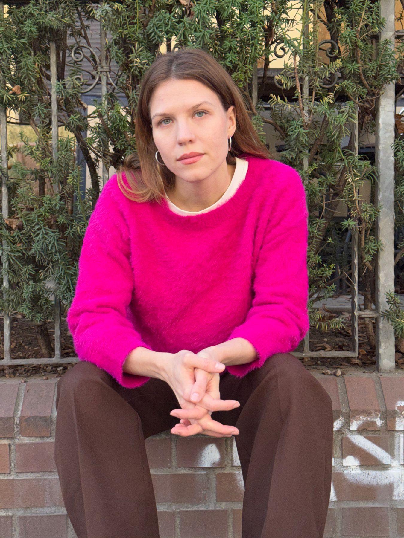 A woman wearing brown trousers and a pink jumper sitting on a wall with her hands clasped