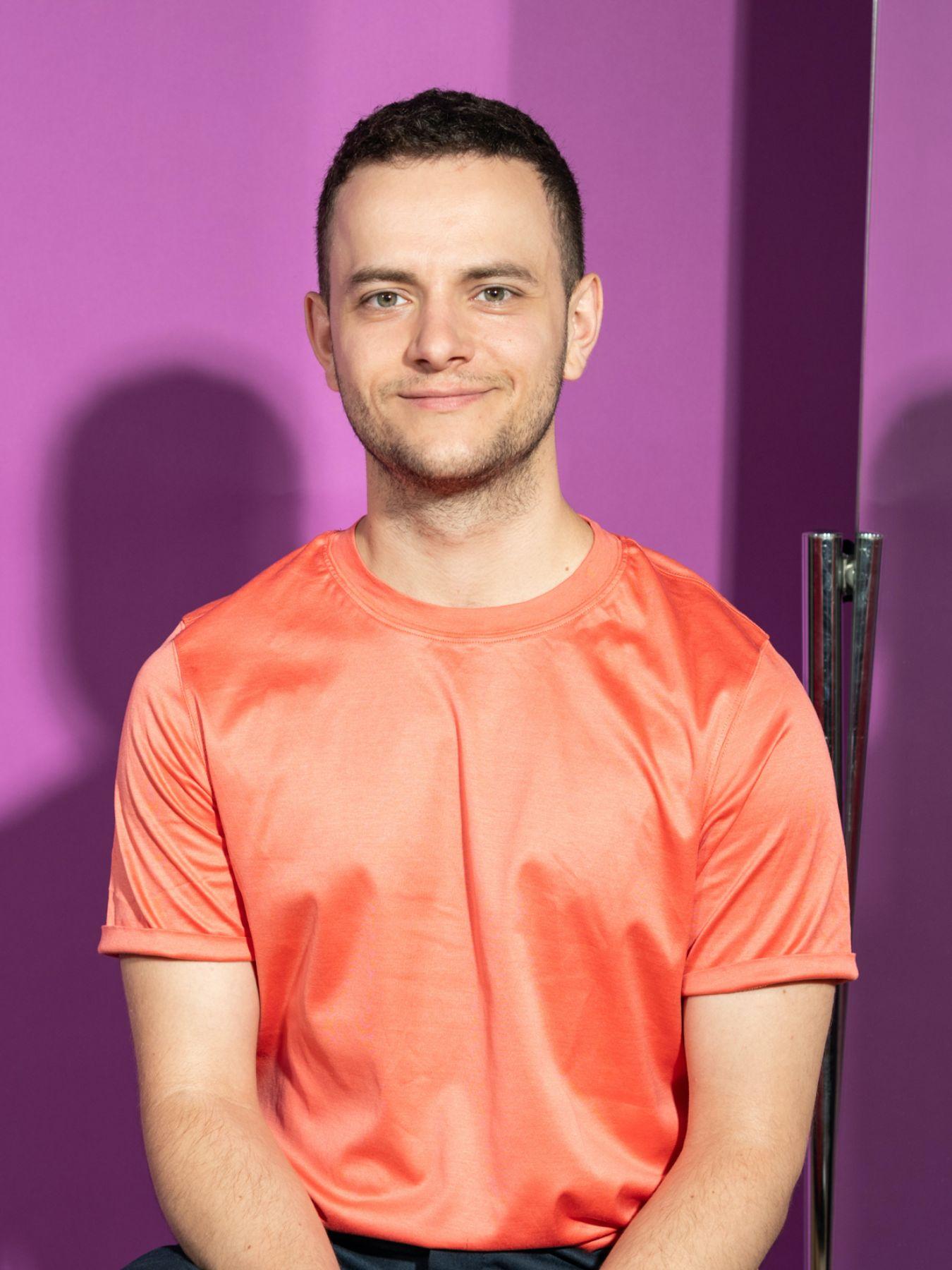 A man with short brown hair sitting in a shiny orange T-shirt in front of a purple wall