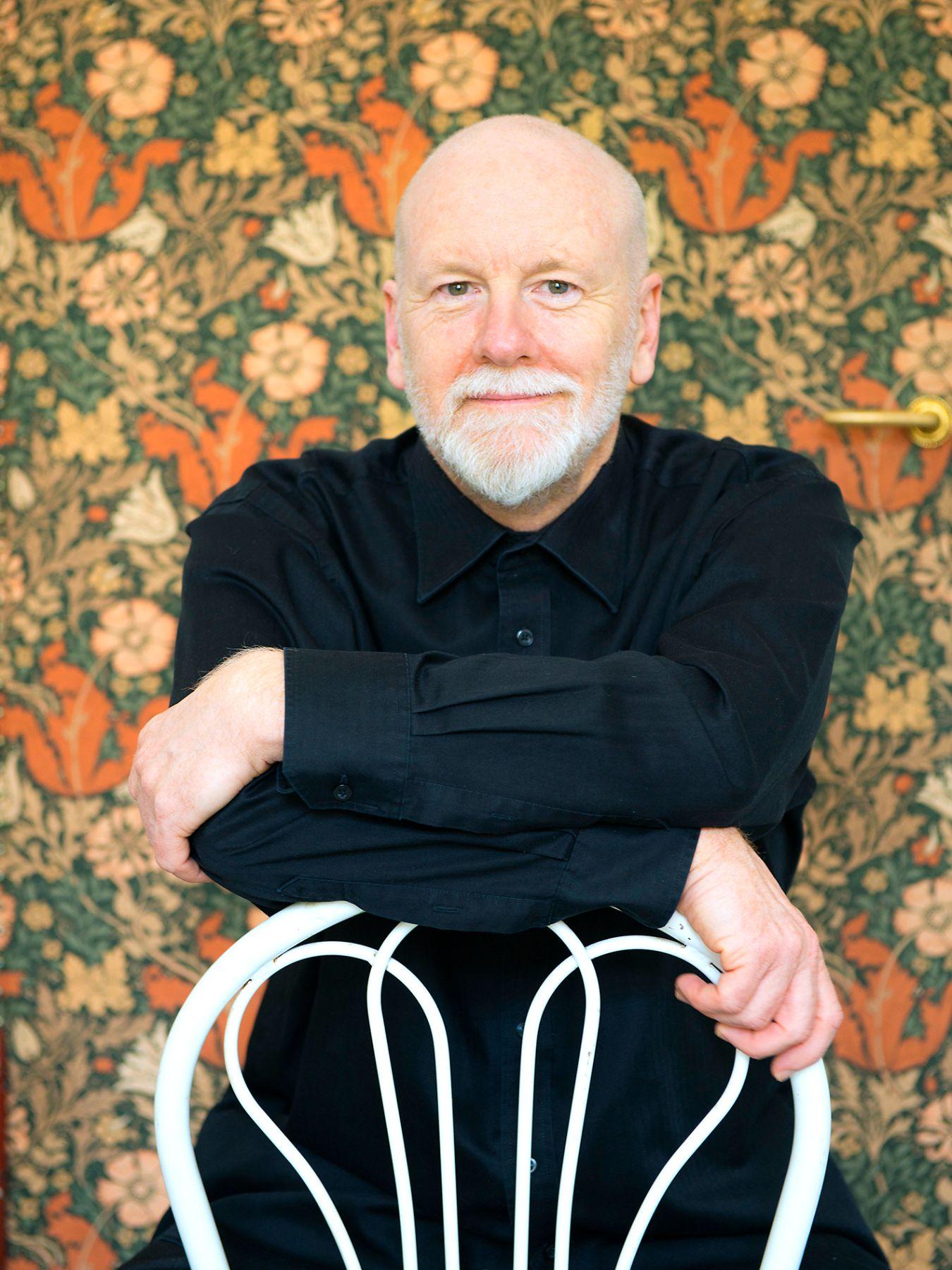 Ein Mann mit weißem kurzen Bart lehnt auf einer Stuhllehne und schaut lächelnd in die Kamera. Im Hintergrund ist eine bunte Blumentapete zu sehen.