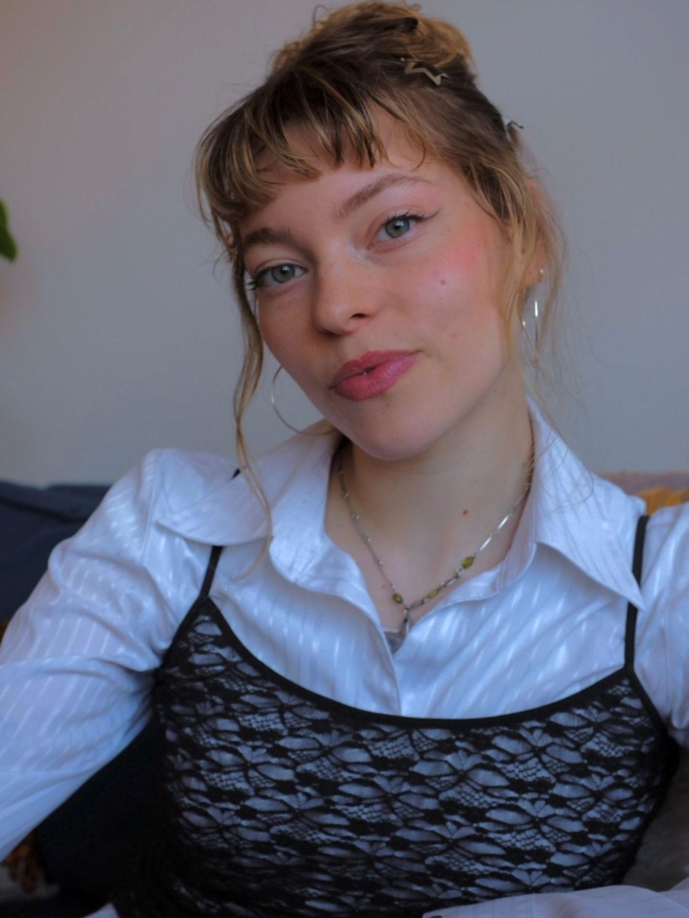 Eine Person mit blondem Haar sitzt auf einem Sofa. Die Person trägt eine weiße Bluse, darüber ein schwarzes Top. Im Hintergrund sind eine Pflanze und eine Diskokugel zu sehen.