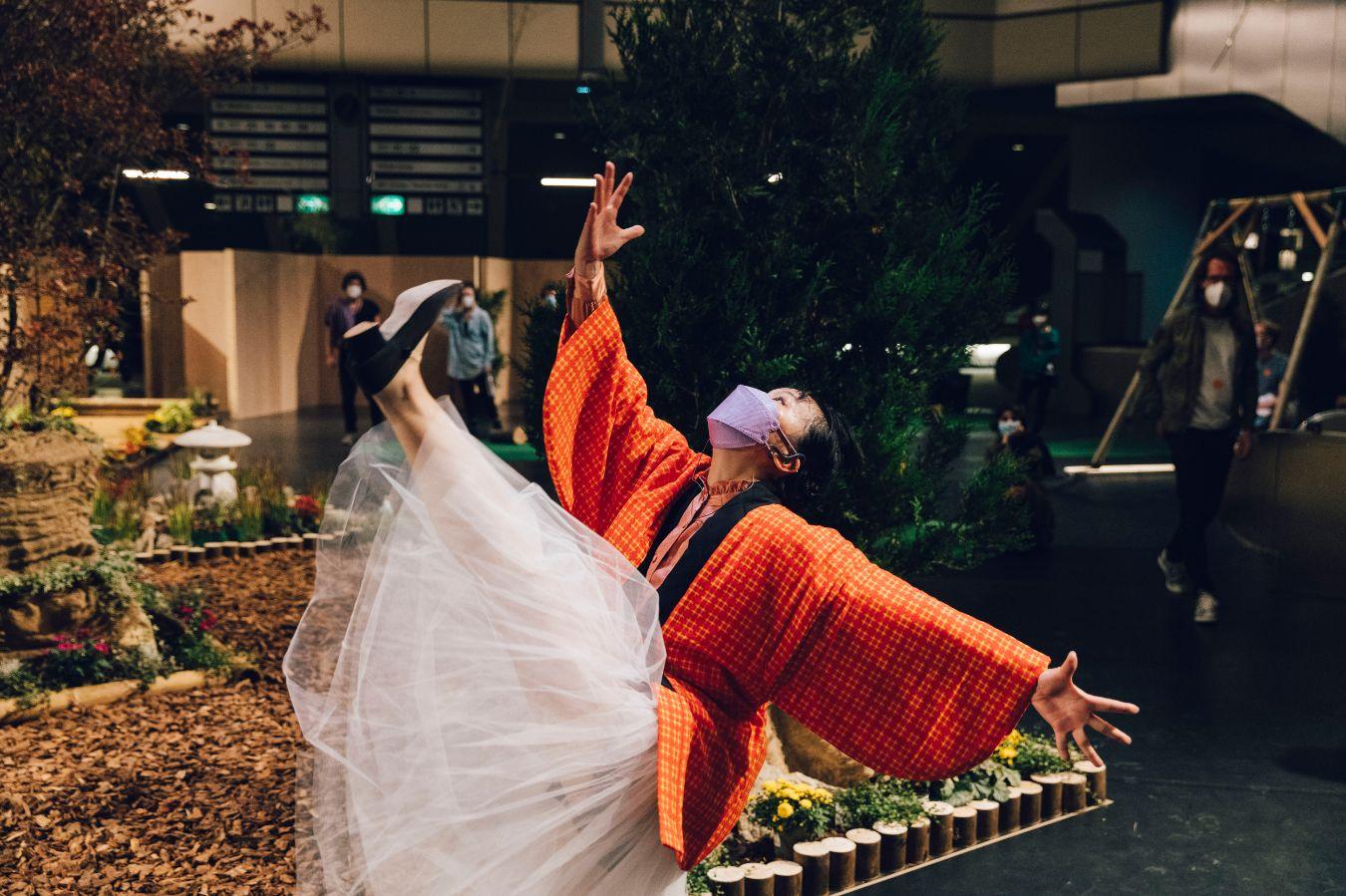 Eine weiblich gelesene Person im weißen Tüllrock und roten Kimono, die ihr linkes Bein in der Luft hält und ihre Arme weit auseinanderstreckt. Sie ist vor einer Gartenlandschaft, die sich in einer Halle befindet.