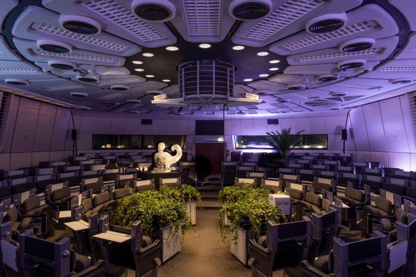 A round, seated hall that reminds one of the interior of a spaceship. The tables in the centre are decorated with green plants. In the back oft he left half of the image, there is a white sculpture.