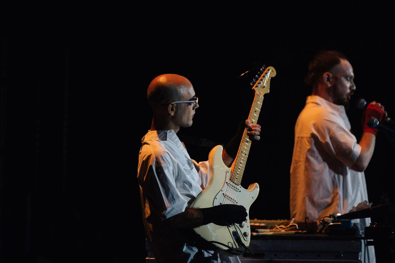 Two male read persons, all dressed in white, during a musical performance. The person in the foreground plays the guitar, the person in the background holds a microphone.