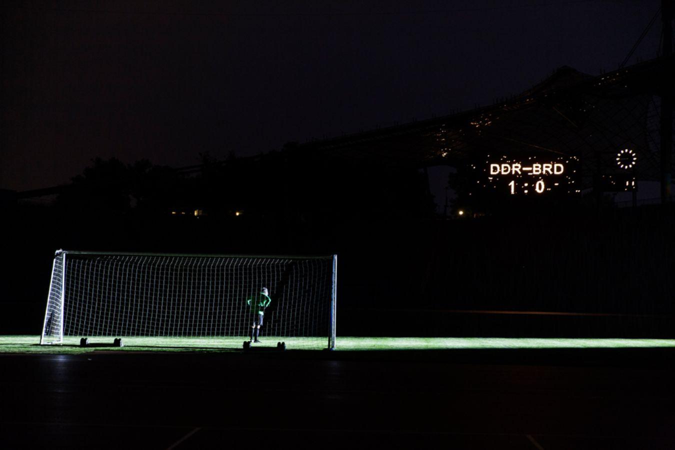 A football goal on a dark pitch. An illuminated sign shows West – East Germany 1:0.