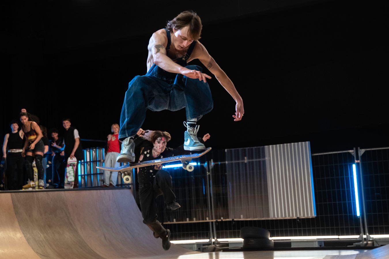 Eine Person mit einem Skateboard in einem Sprung auf einer Half-Pipe. Sie wird von anderen Skatern dabei beobachtet.