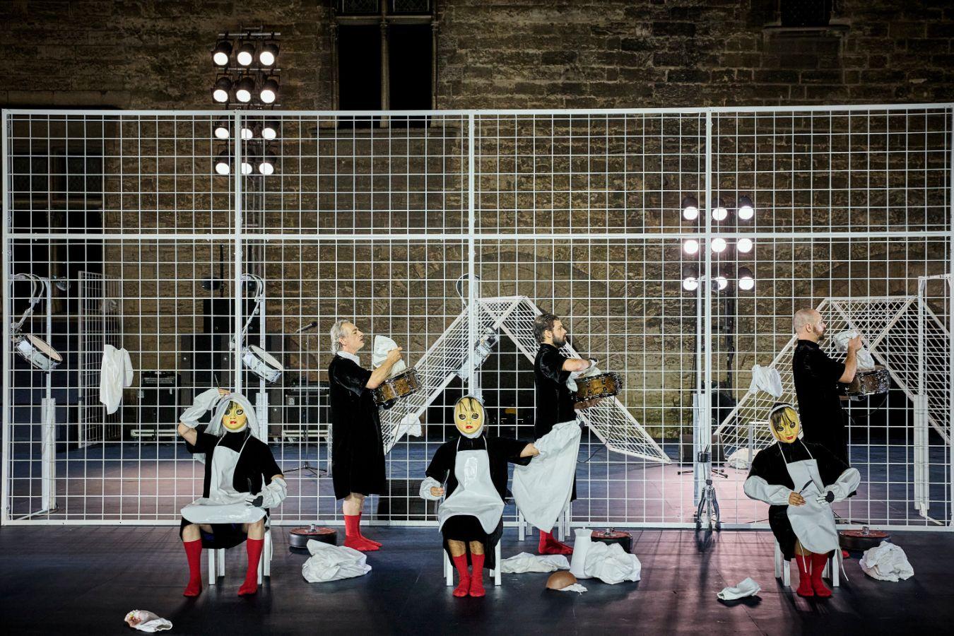 Performer*innen mit Puppen-haften Masken vor einem Gitterzaun