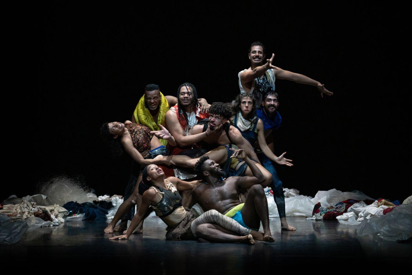 A group of dancers sit and stand close together surrounded by discarded clothes.