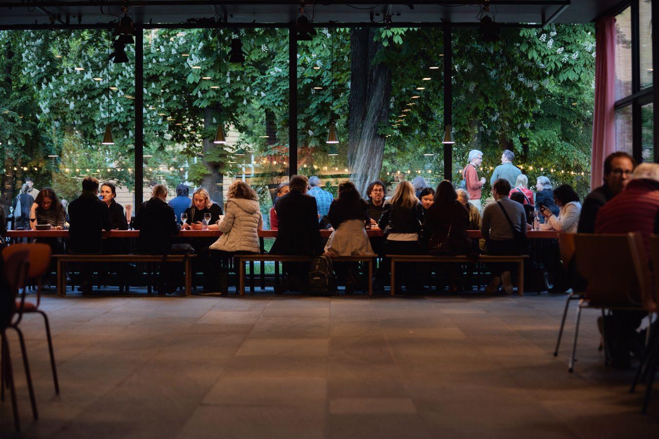 People sitting at long wooden tables in a room with large glass fronts, green trees visible outside.