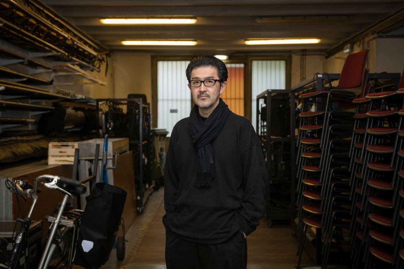 A man standing in a narrow storage room filled with stacked chairs, equipment racks and a bicycle, facing the camera.
