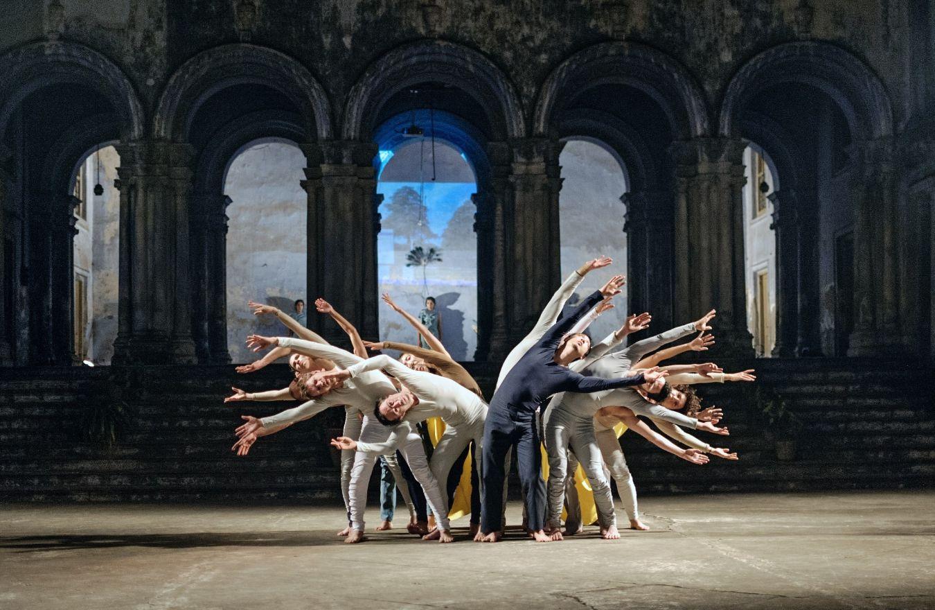 Tanzensemble in synchron gebogener Pose in einer historischen Halle mit beleuchtetem Hintergrund.
