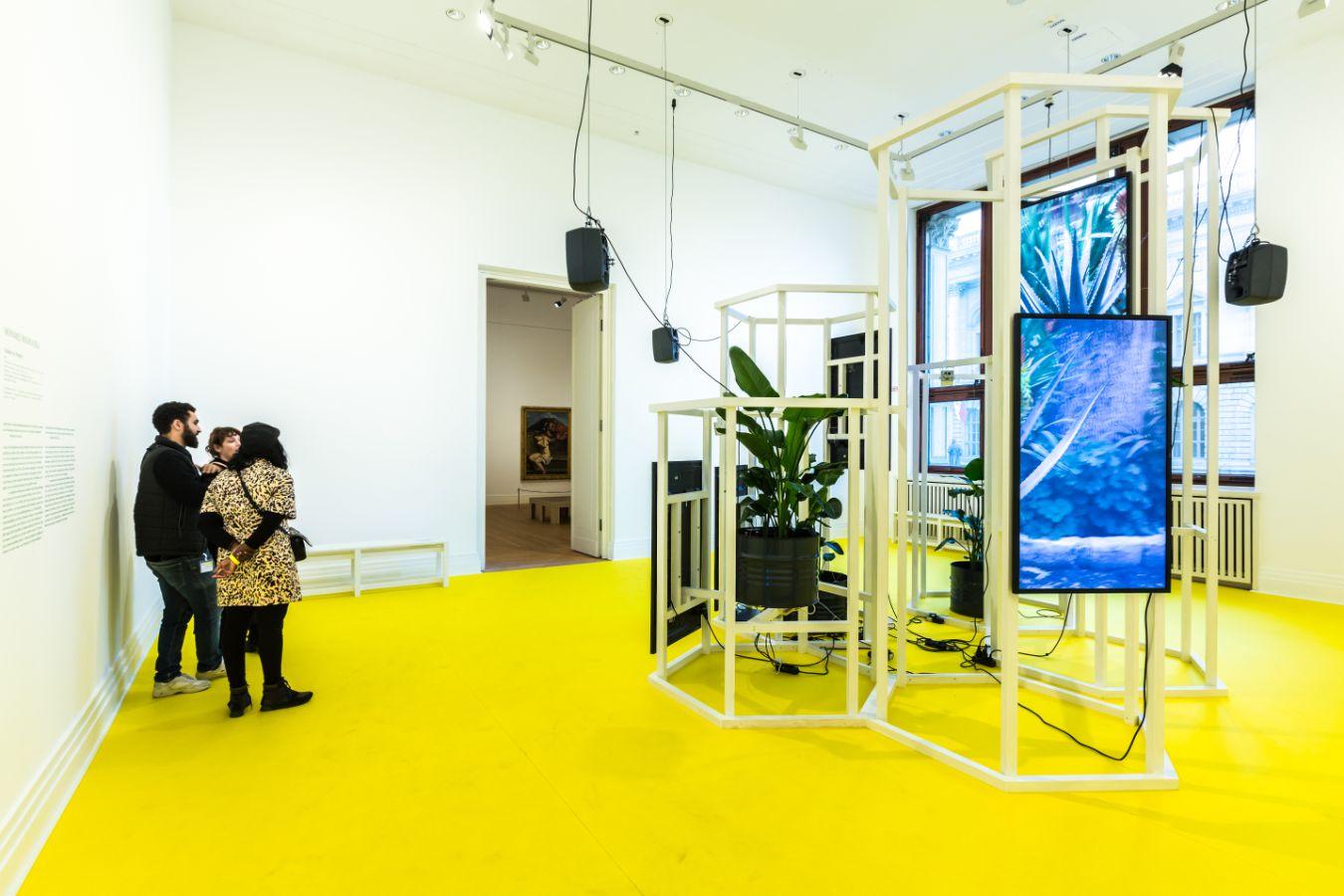 Das Foto zeigt einen belebten Ausstellungsraum mit der Installation, bestehend aus einem gelben Bode, einer Holzkonstruktion, Bildschirmen, Lautsprechern und Topfpflanzen.