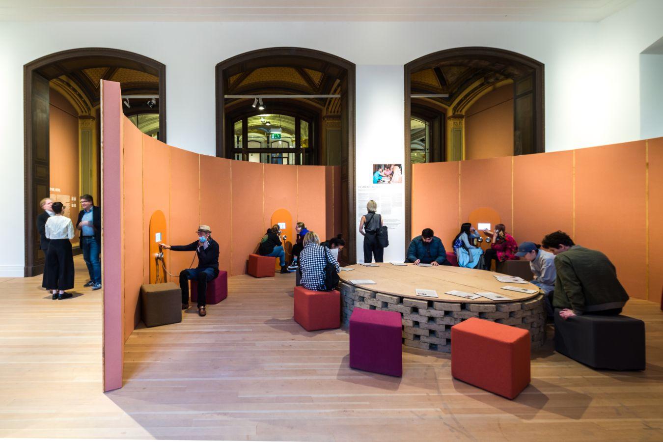 Eine belebte Installationsansicht mit einer orangenen Wand, die in einem Halbkreis aufgestellt ist mit Hörvorrichtung. Davor befindet sich ein Tisch auf rund angeordneten Lehmziegeln und Sitzmöglichkeiten.
