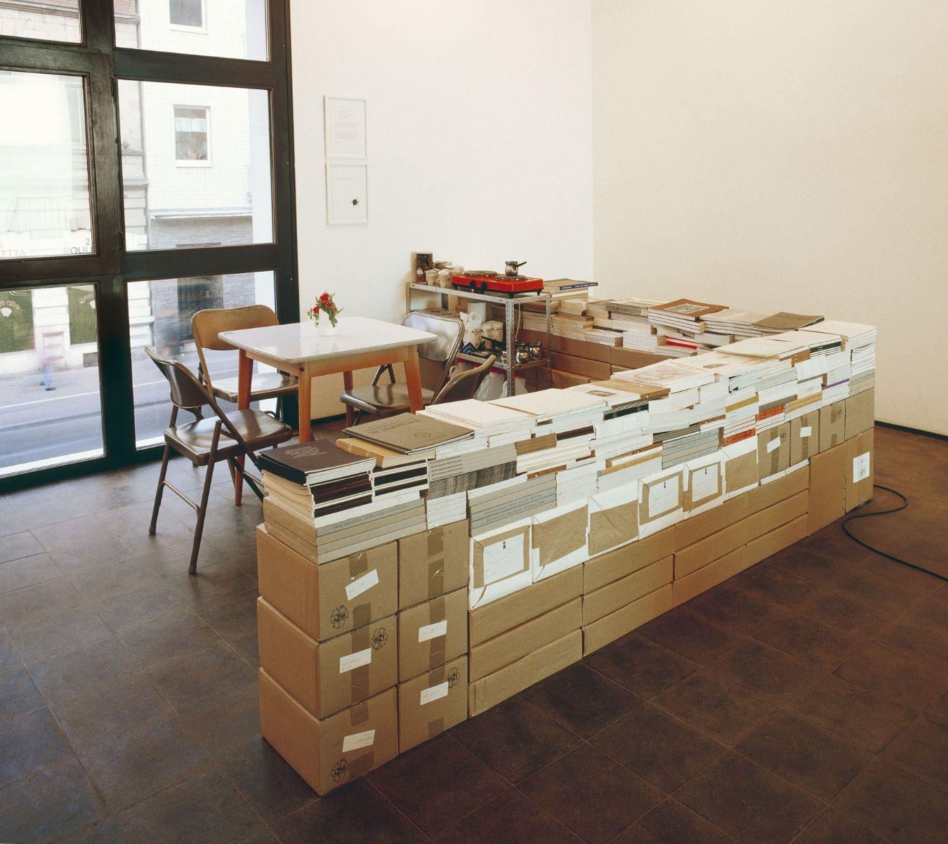 Ansicht von Rirkrit Tiravanijas Installation "untitled 1993 (café deutschland)", ein Tisch mit Stühlen vor einem Fenster. Um den Tisch stehen Kartons auf denen Papierkataloge liegen.