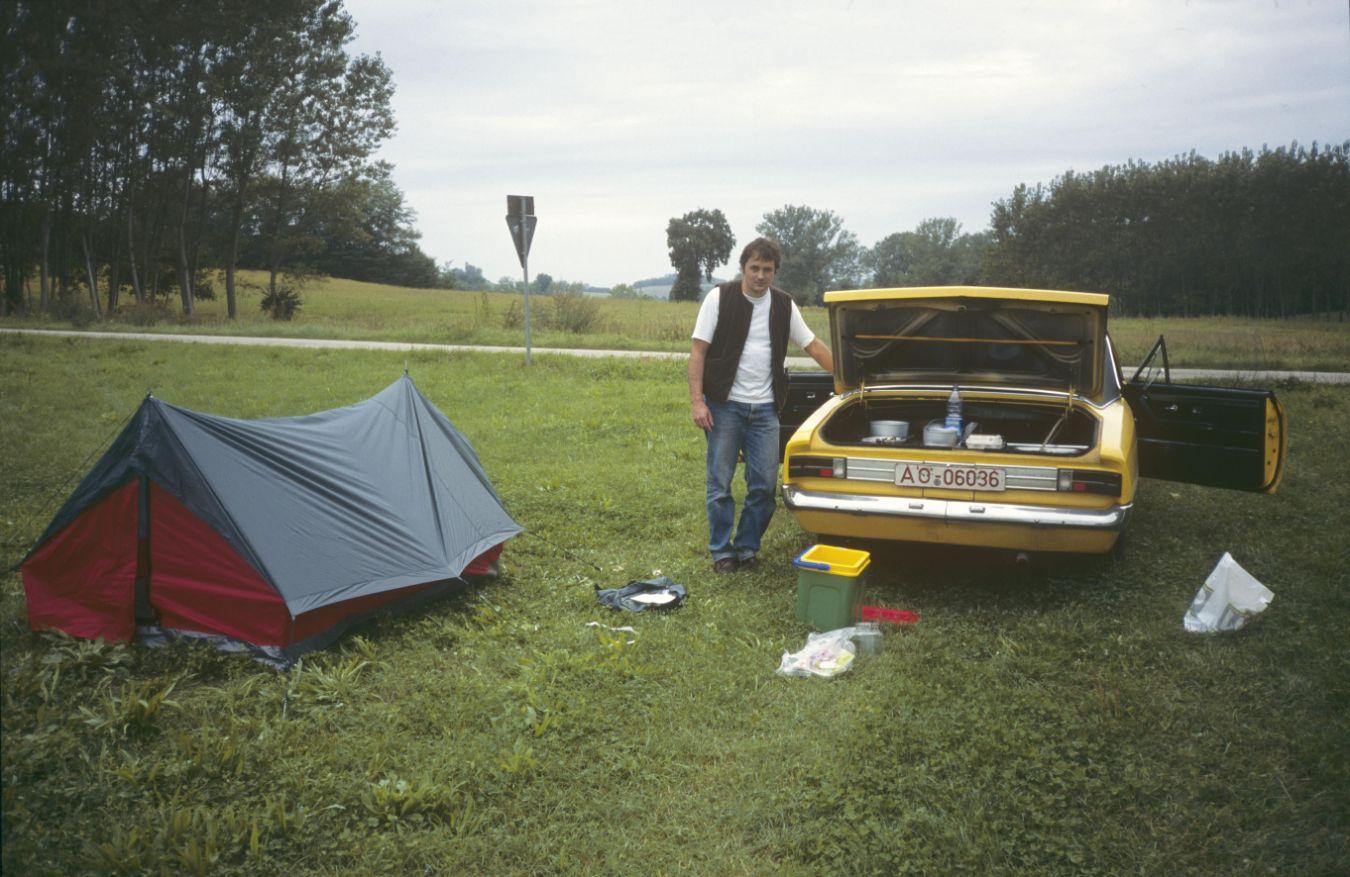 Ein Foto von einem gelben Auto, das auf einer Wiese steht. An dem Auto lehnt ein Mensch und schaut Richtung Kamera. Neben dem Menschen ist ein Zelt aufgeschlagen.