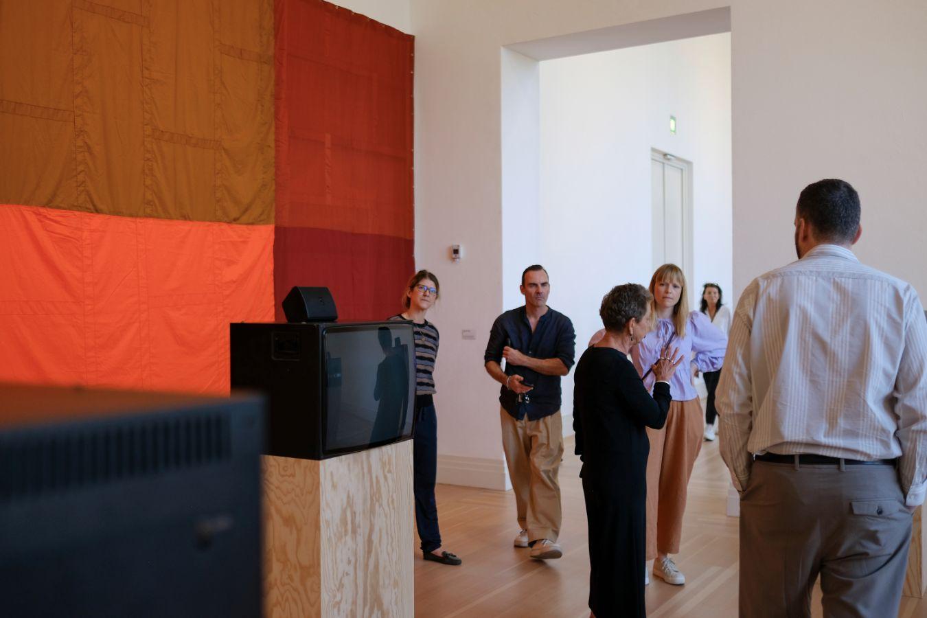 Several people are standing in an exhibition room listening to a guided tour.