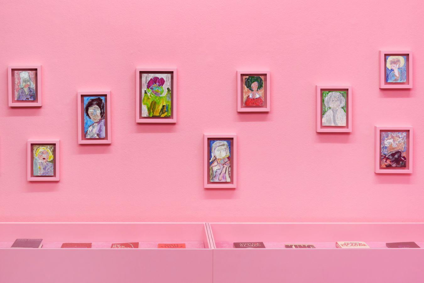 The picture shows a pink wall with different-sized framed pictures hanging on it.