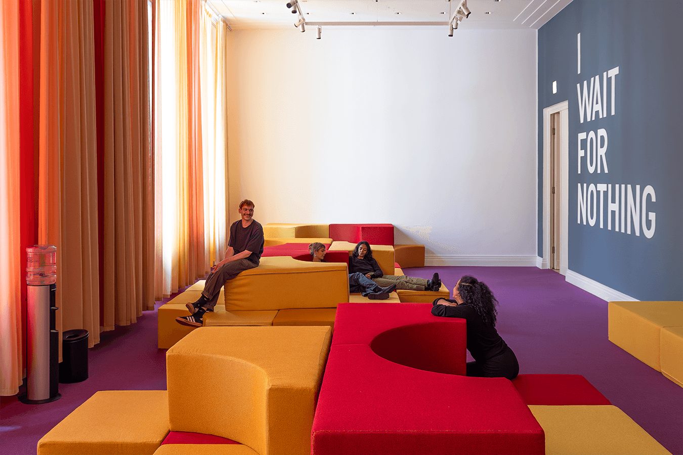 Eine Aufnahme von einem Raum mit lila Teppichboden, in dem sich verschiedene Polsterelemente in gelb und rot befinden, darauf sitzend vier Personen. Über die Länge der linken Wand ziehen sich gelbe und rote Vorhänge, die rechte Wand ist petrolfarben mit eine großen Schriftzug in weiß "I WAIT FOR NOTHING".