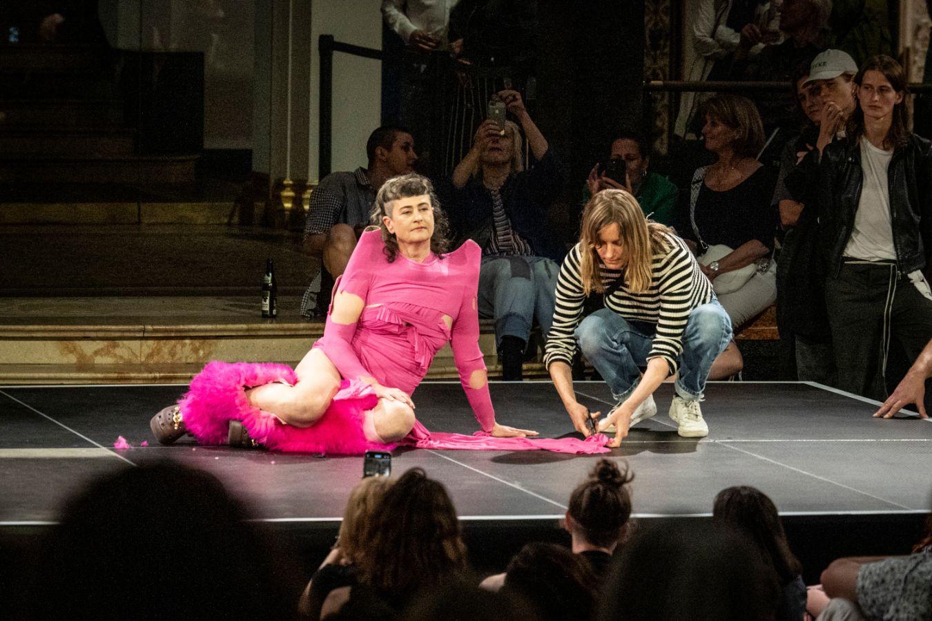 A person wearing pink clothing sitting on a stage, with another person to their right cutting a piece of the pink clothing with scissors.