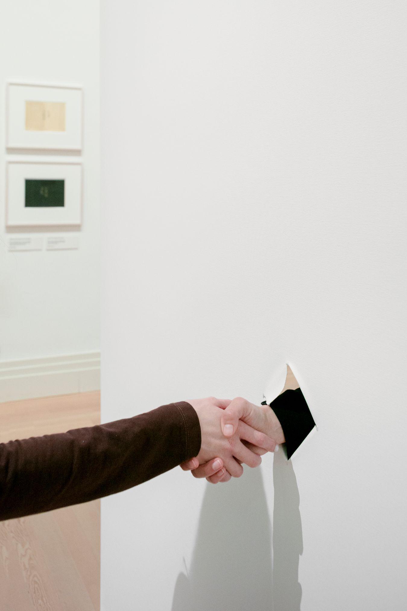 Through a hole in a white screen, two people who cannot be seen shake hands.
