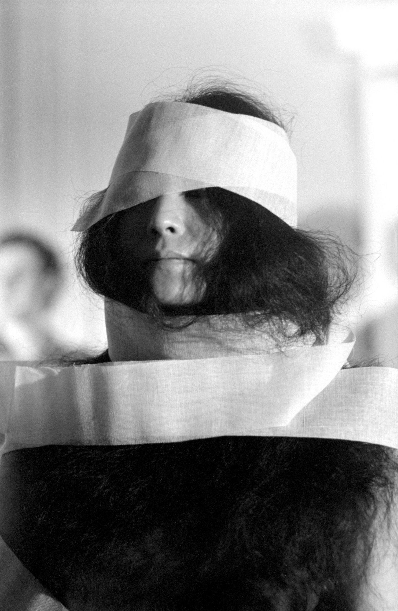 A black-and-white portrait of Yoko Ono with a white bandage wrapped around her head, eyes, and neck.