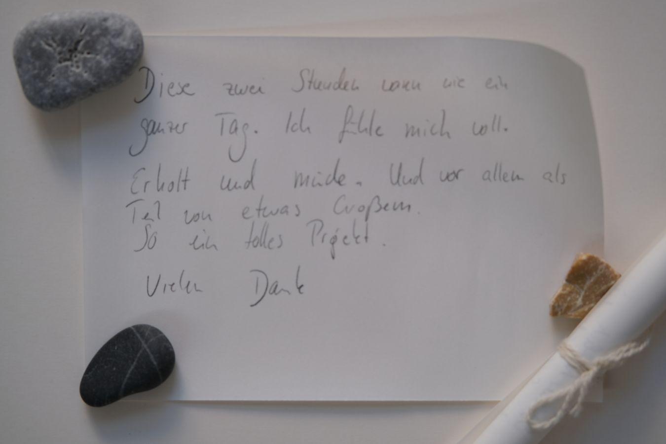 Handschriftlicher Text auf Papier mit Steinen und gerolltem Papier. Textinhalt: ‚Diese zwei Stunden waren wie ein ganzer Tag. Ich fühle mich wohl. Erfüllt und müde. Und vor allem als Teil von etwas Großem. So ein tolles Projekt. Viele Dank.‘