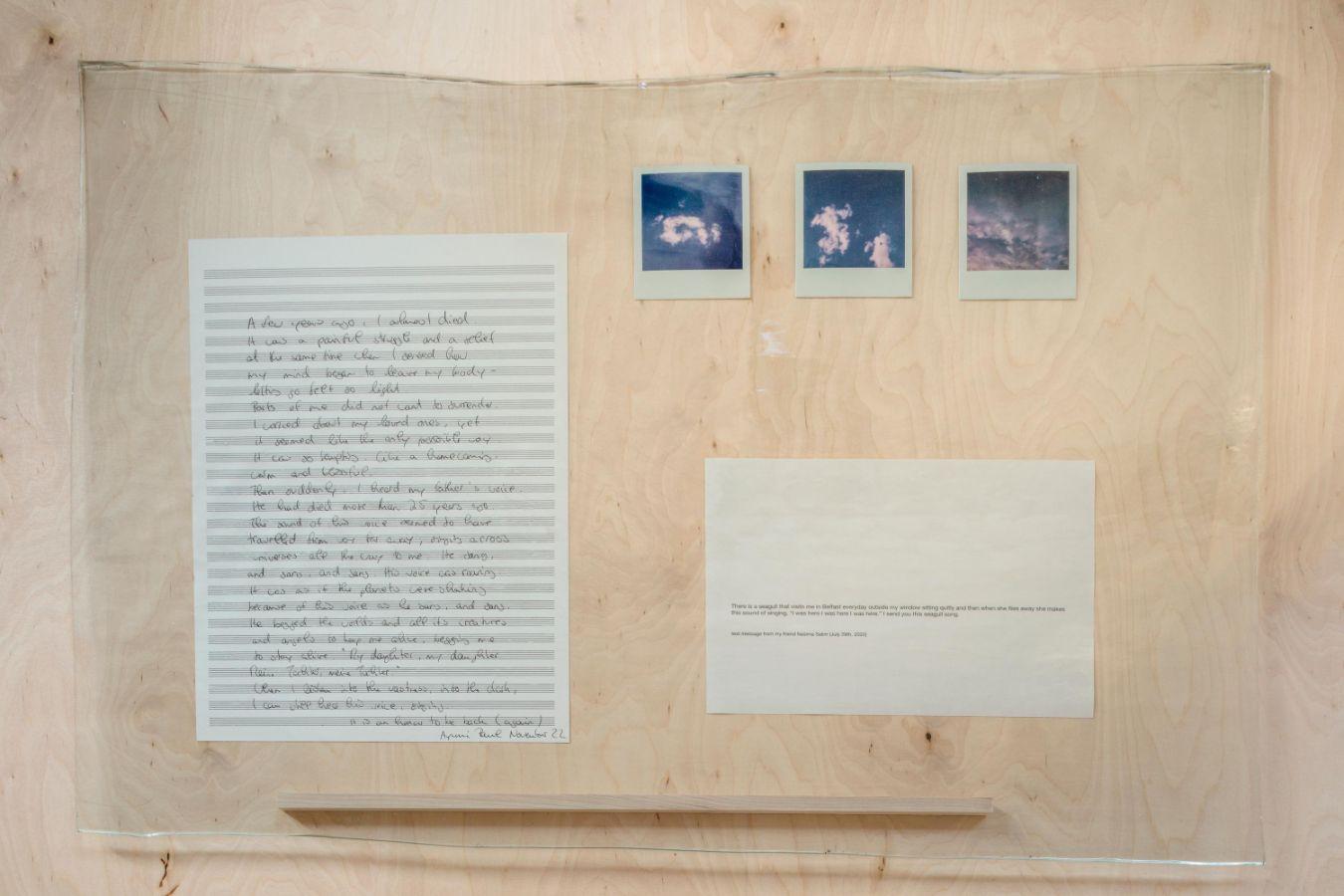 Auf hellem Holz montierte Glasplatte mit Polaroidbildern von Wolken, handgeschriebenen Notizen und einem bedruckten Blatt.
