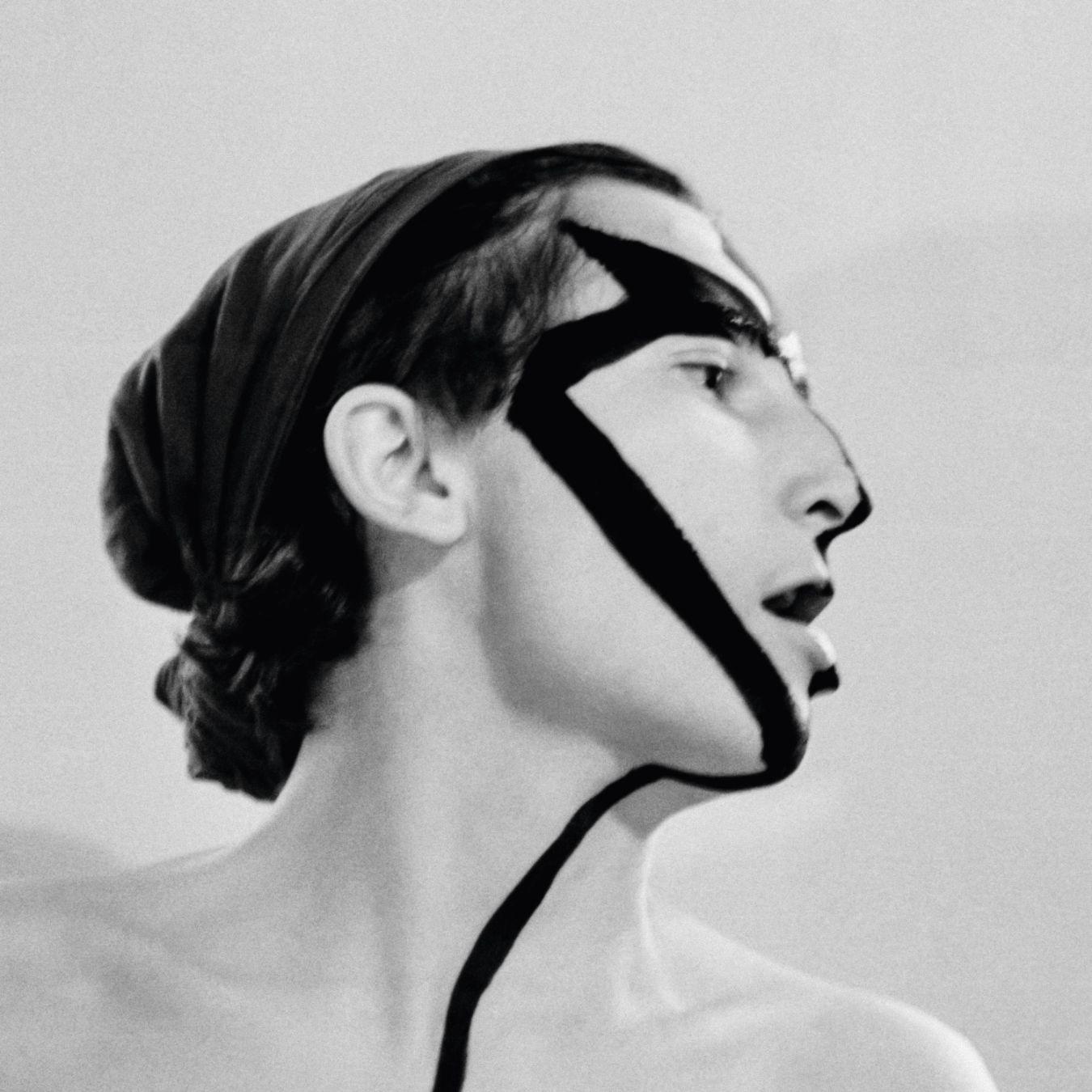 A black‑and‑white profile portrait of a person with the head slightly tilted upward. Wide, dark paint stripes run diagonally across the face and down the neck, crossing the forehead, eye area, and cheek.