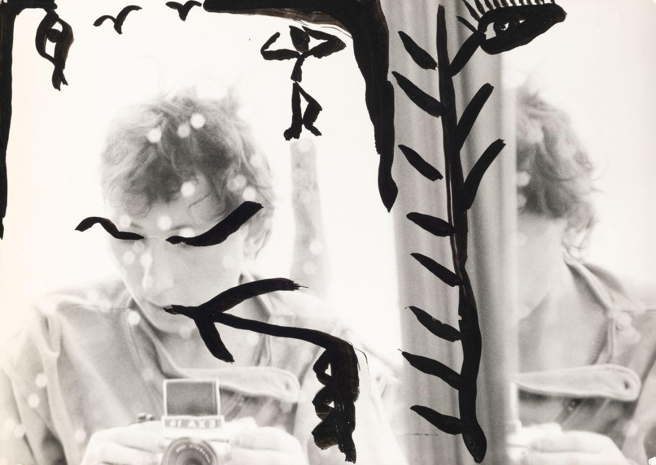 Ein mit schwarzen Mustern bemaltes Selbstporträt von Gabriele Stötzer, die sich mit einer Analogkamera im Spiegel fotografiert.